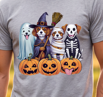 A close-up of a heather gray t-shirt featuring a colorful graphic of four dogs dressed in Halloween costumes (ghost, witch, mummy, skeleton) sitting behind three jack-o'-lantern pumpkins.