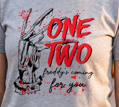 A close-up of a heather gray t-shirt featuring a graphic print of a clawed hand holding a knife, with the text 'ONE TWO' in large red brush lettering and 'freddy's coming for you' in black script below.
