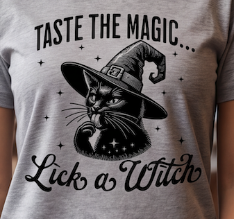 A close-up of a light heather gray t-shirt featuring a black graphic of a witch cat wearing a pointed hat, with the text 'TASTE THE MAGIC.