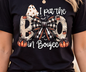 A close-up of a person wearing a black graphic T-shirt with a Halloween-themed design. The shirt features the phrase 'I put the BOO in Boujee' in white script and block lettering, decorated with a large black-and-white buffalo plaid bow, white ghosts, and orange pumpkins.