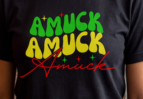 A close-up of a person wearing a black t-shirt with a colorful graphic print that reads 'AMUCK AMUCK Amuck' in green, yellow, and red retro-style lettering with small star accents.