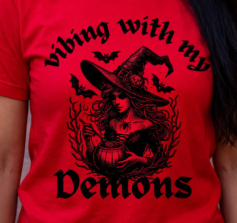 A close-up of a person wearing a bright red T-shirt with a large black graphic print. The design shows a witch in a pointed hat holding a cauldron, surrounded by bats and the text 'vibing with my Demons' in a gothic font.
