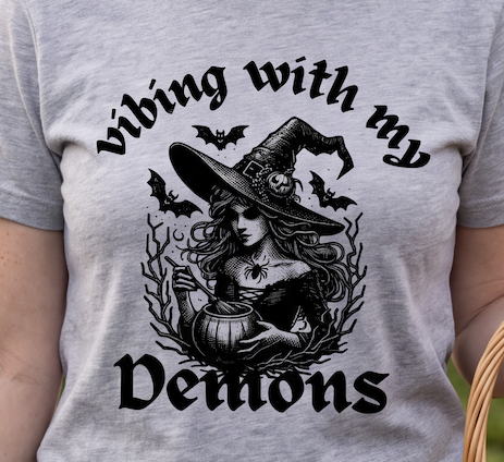 A close-up of a person wearing a heather gray t-shirt with a black graphic print of a witch, bats, and the text 'vibing with my demons'.