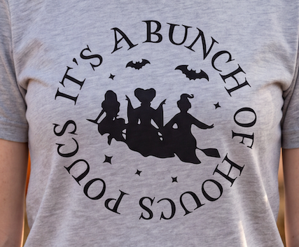 A close-up of a person wearing a heather gray t-shirt with a black graphic print that reads 'IT'S A BUNCH OF HOCUS POCUS' around a silhouette of three witches riding broomsticks.