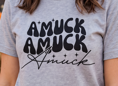 A close-up of a person wearing a heather gray t-shirt with a black graphic text design that reads 'AMUCK AMUCK Amuck' with small star accents.