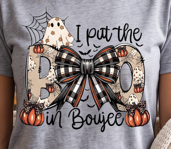 A close-up of a person wearing a heather gray t-shirt with a graphic print that reads 'I put the B in Boujee' in black script. The graphic features a large bow made of black, white, and orange plaid, flanked by two patterned 'B' letters decorated with pumpkins, bats, and a spiderweb, with a white ghost on the upper left.