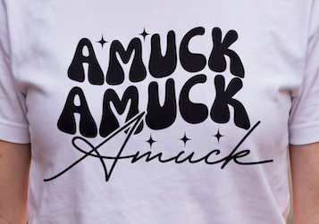 A close-up of a person wearing a white T-shirt with a bold black graphic print that reads 'AMUCK AMUCK Amuck' in stylized lettering.