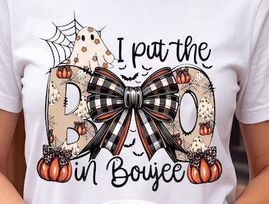A close-up of a person wearing a white T-shirt with a large Halloween graphic. The design features the phrase 'I put the BOO in Boujee' in black script, with the letters filled with tan animal-print patterns, orange pumpkins, black spiderwebs, and a large black-and-white plaid bow in the center.