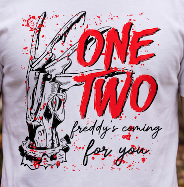 A close-up of a white T-shirt featuring a horror graphic print. The design shows a black-and-white illustrated clawed hand holding a gun, with bold red text reading 'ONE TWO' and smaller black script text reading 'freddy's coming for you.