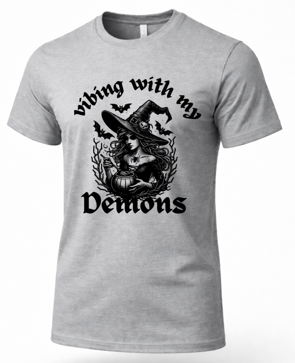 A heather gray short-sleeve crewneck t-shirt featuring a black graphic print of a witch sitting in a pumpkin with bats flying around her, surrounded by the text 'Vibing with my Demons'.