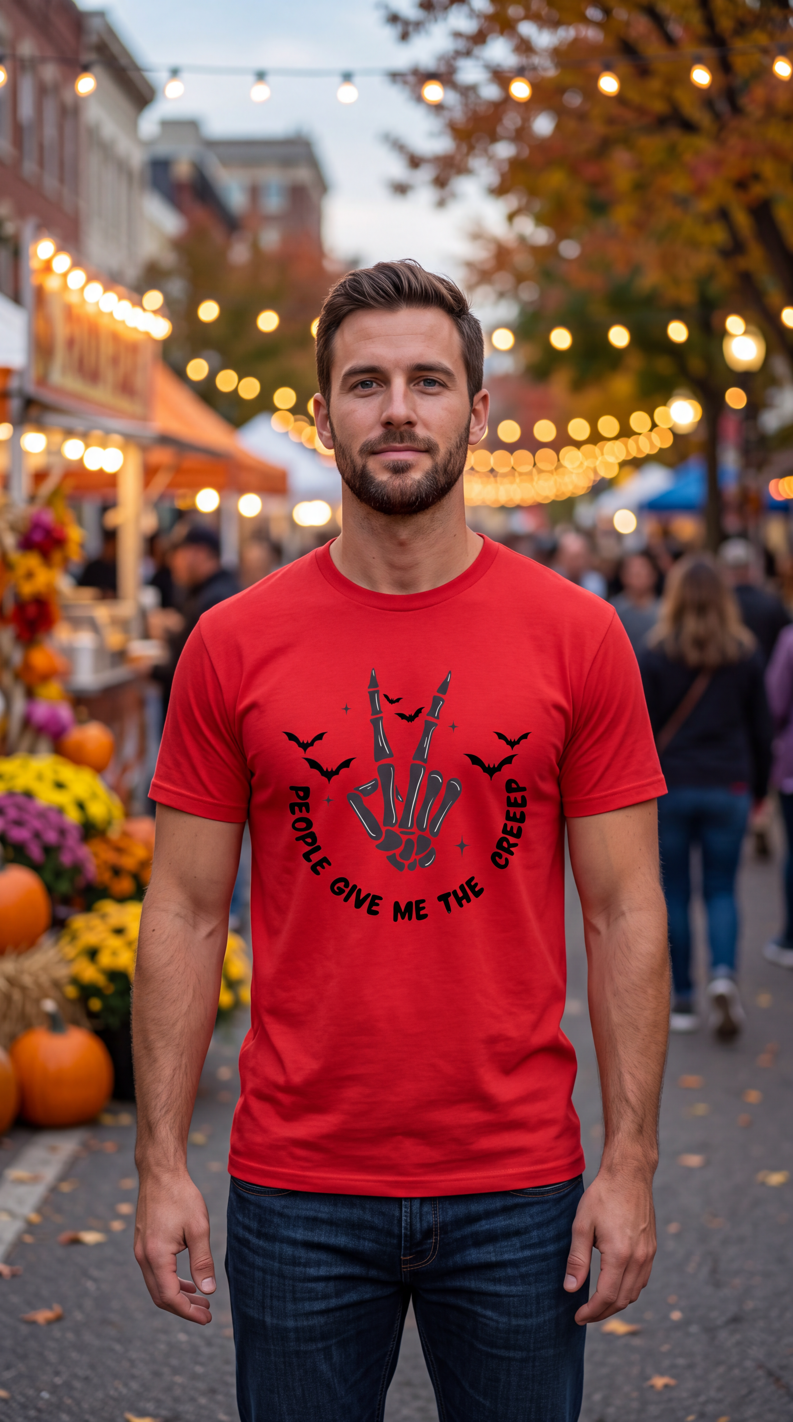 A man wearing a bright red graphic t-shirt with a black skeleton hand making a peace sign and the text 'PEOPLE GIVE ME THE GREEK' stands in the center of an outdoor autumn street market with warm string lights and pumpkins in the background.