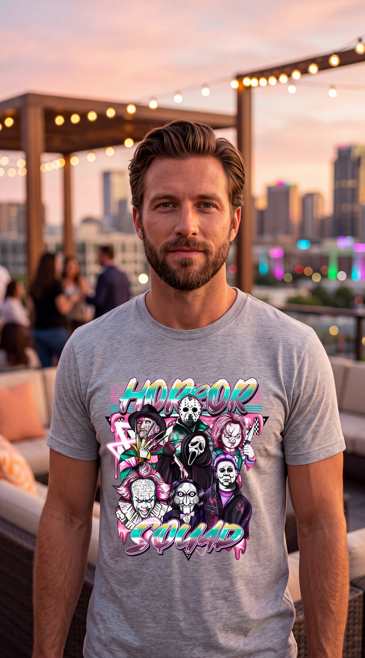 A man wearing a heather gray t-shirt with a colorful graphic print featuring the text 'HORROR SQAUD' and stylized horror character illustrations, standing on a rooftop patio at sunset.