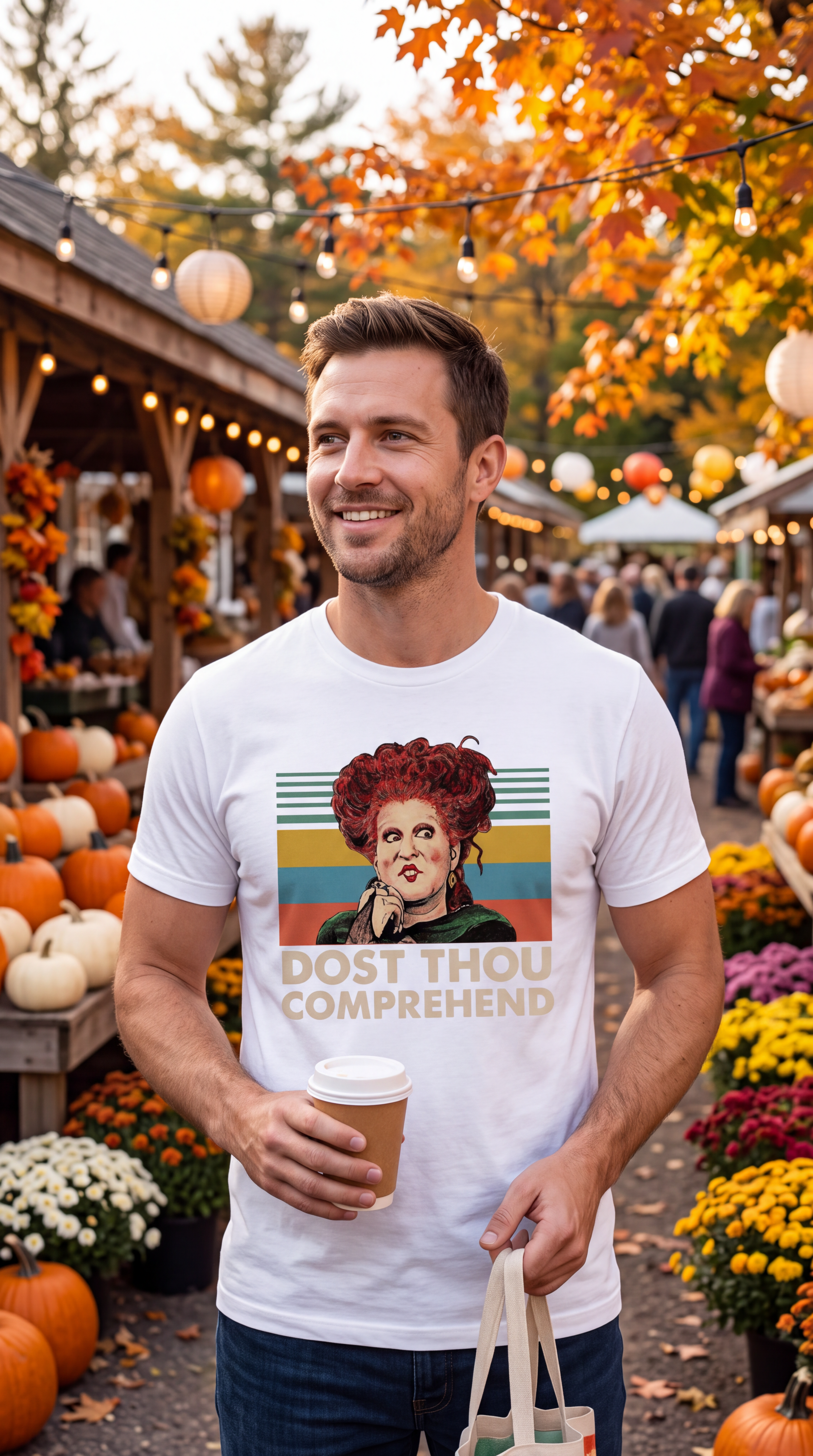 A man with curly red hair stands on a brick street wearing a black graphic T-shirt featuring a retro-style illustration of a woman with red hair and the text 'DOST THOU COMPREHEND'. The background shows a cozy, autumnal street scene with pumpkins, fall leaves, and warm string lights.
