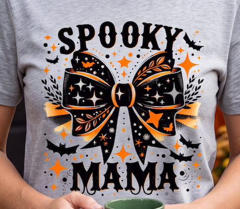 A person wearing a heather gray graphic T-shirt with a large black and orange bow design and the words 'SPOOKY MAMA' on the front, holding a green mug. The photo is cropped from the chest down with the person's arms visible.