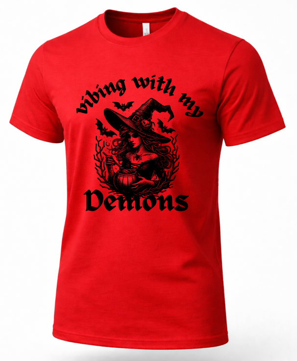 A red short-sleeve crewneck t-shirt featuring a black graphic print of a witch sitting in a pumpkin with bats flying around her, surrounded by the text 'Vibing with my Demons'.