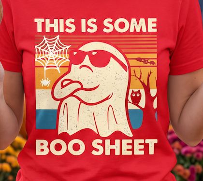 A red close up short-sleeve graphic T-shirt worn by a person, featuring a retro-style cream ghost illustration with sunglasses, a spiderweb, owl, and trees over a sunset background, with the text 'THIS IS SOME BOO SHEET'.