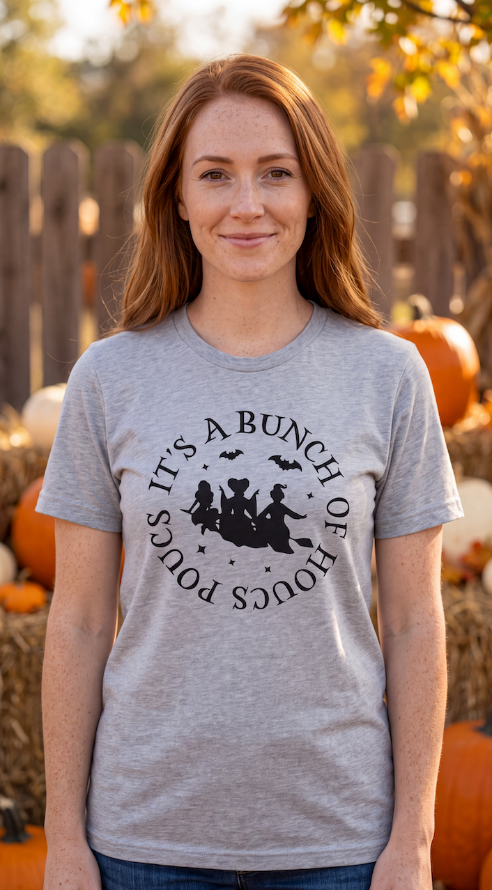 A smiling woman stands in front of a pumpkin patch, wearing a heather grey short-sleeve T-shirt with a black circular graphic and text reading 'IT'S A BUNCH OF HOCUS POCUS' around a witch riding a broomstick with bats.