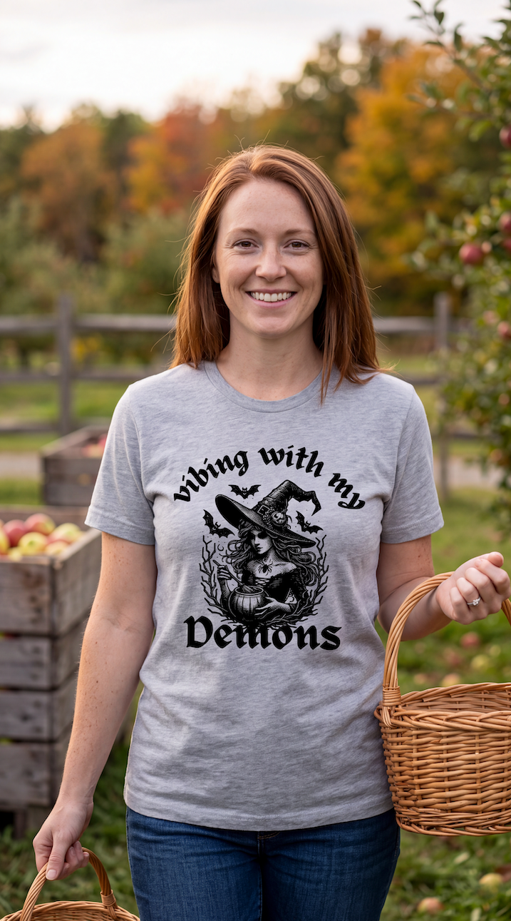 A smiling woman stands outdoors in an apple orchard wearing a heather gray graphic T-shirt with a black witch illustration and the text 'Vibing with my Demons.' She is holding wicker baskets, with apple crates and trees in the background.