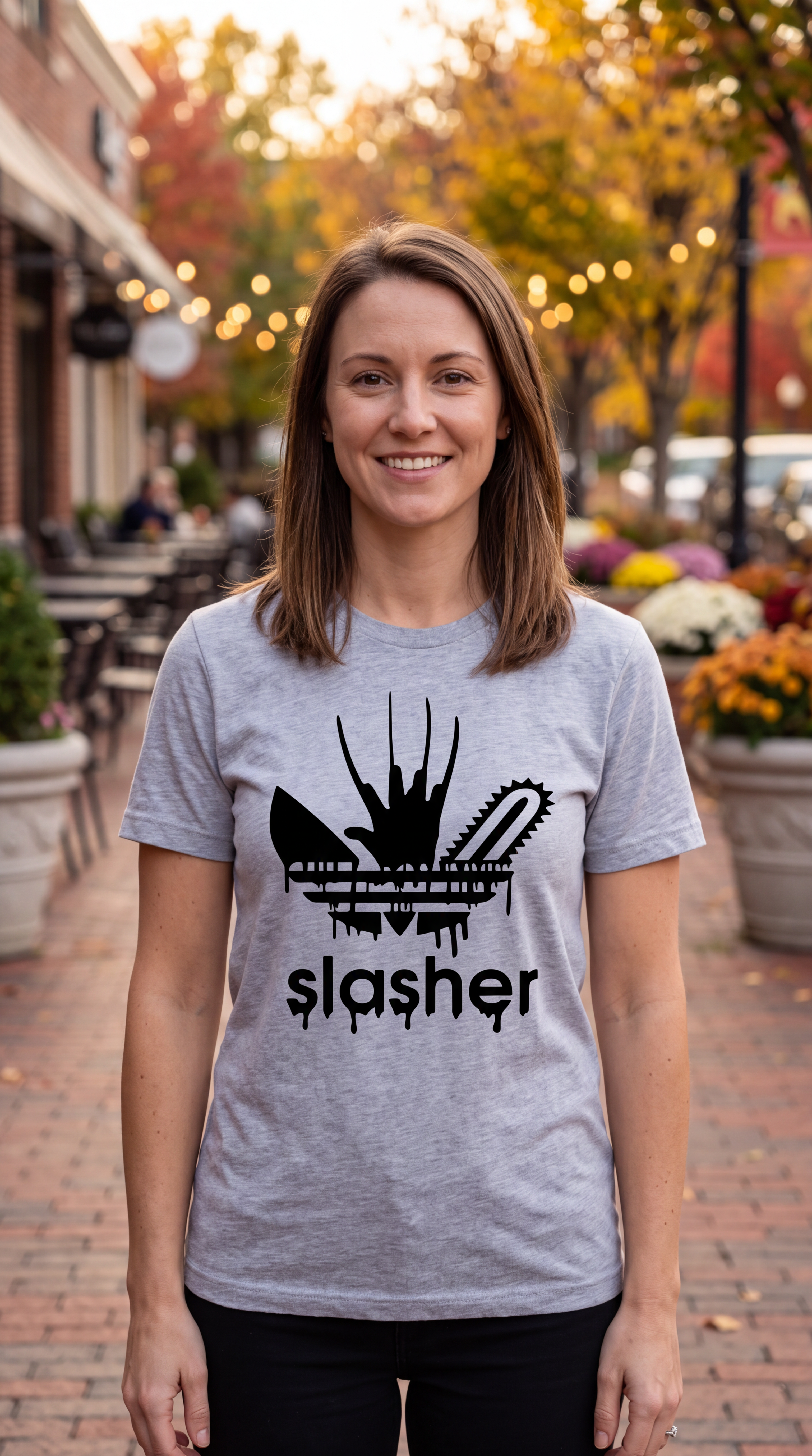 A smiling woman stands outdoors on a brick walkway wearing a light heather gray t-shirt with a black graphic of a chainsaw and the word 'slasher'.