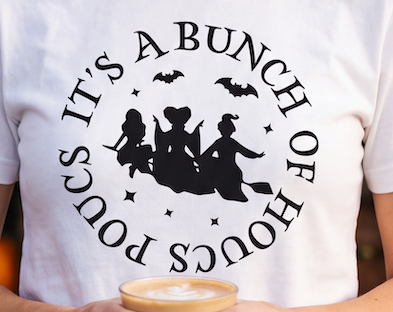 A white short-sleeve T-shirt worn by a person, featuring a black circular graphic with the text 'IT'S A BUNCH OF HOURS POURS' around a silhouette of three witches riding broomsticks with bats and stars.