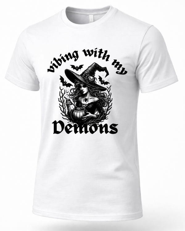 A white short-sleeve crewneck t-shirt with a black graphic print on the front. The graphic features a witch wearing a large pointed hat, surrounded by bats, a pumpkin, and the gothic text 'Vibing with my Demons'.