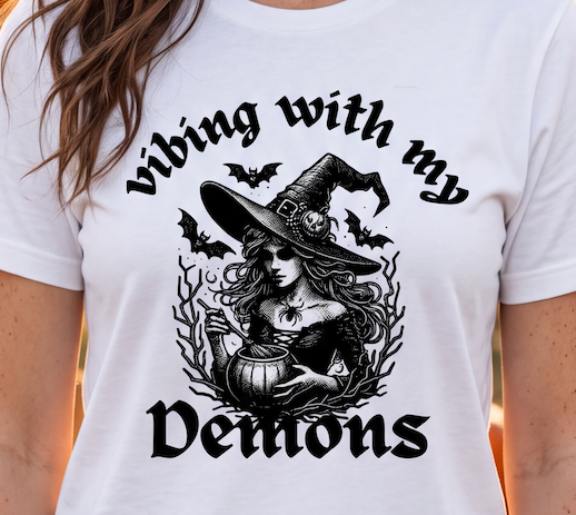 A white short-sleeve graphic T-shirt worn by a model, featuring a black illustration of a witch in a hat holding a cauldron, surrounded by bats and the text 'vibing with my demons'.