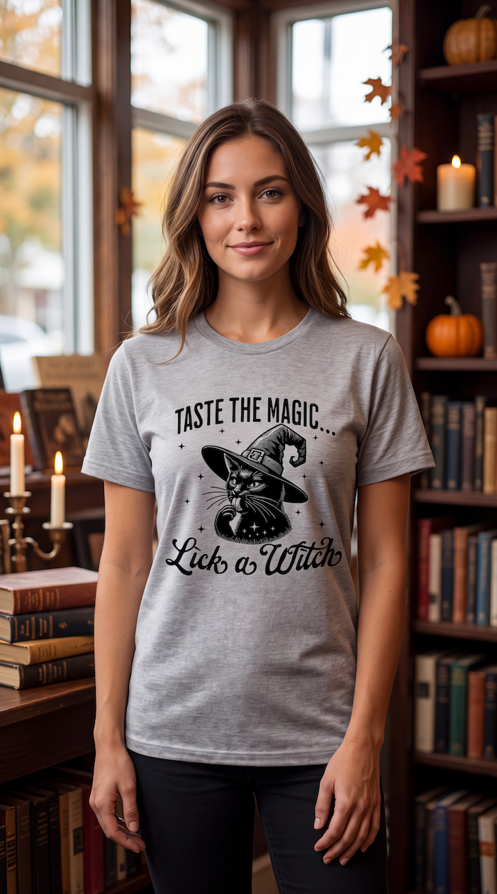 A woman stands in a cozy, book-filled room wearing a heather gray graphic t-shirt with a black print of a witch cat and the text 'TASTE THE MAGIC.