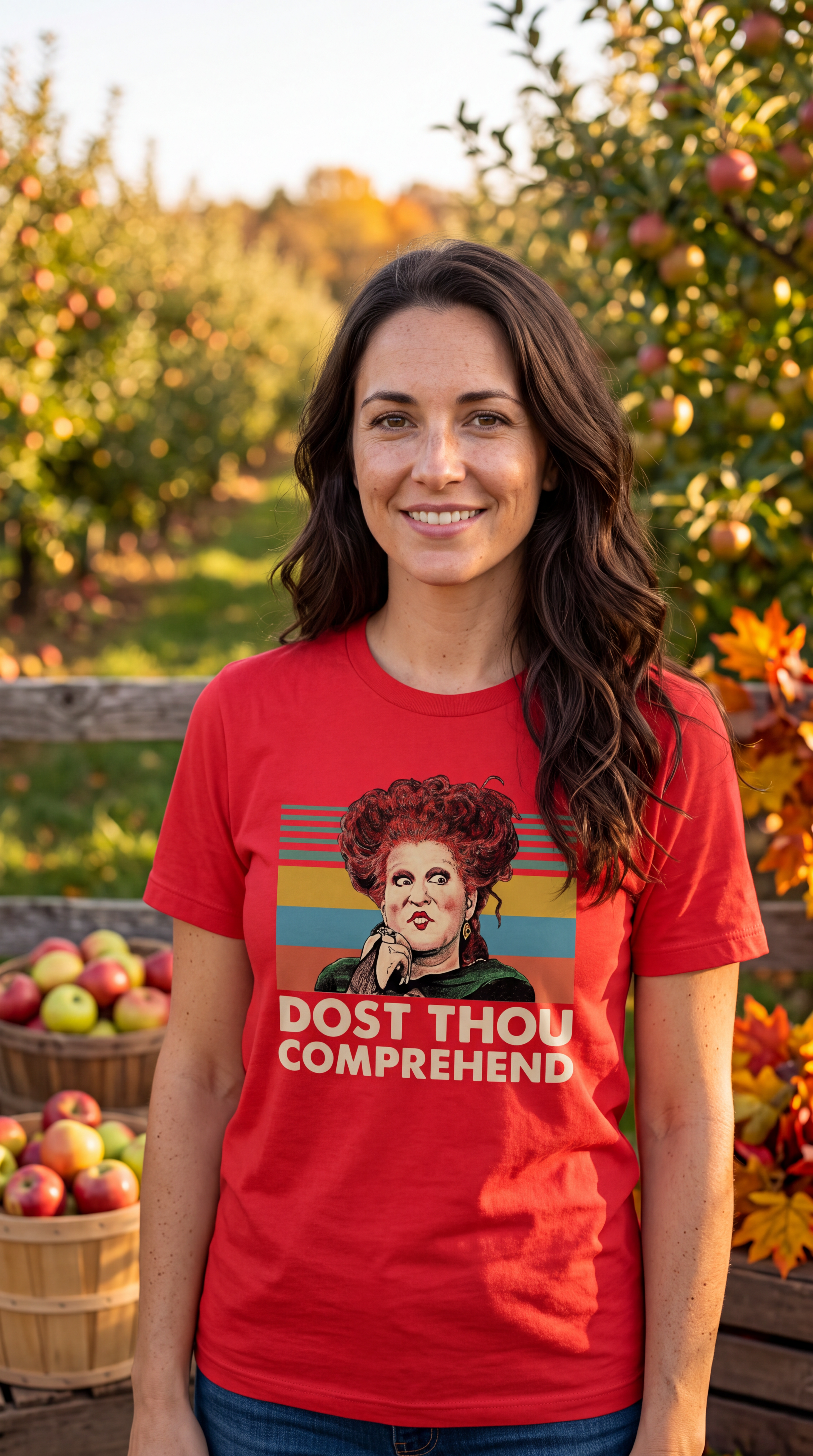 A woman stands in an apple orchard wearing a red graphic T-shirt with a vintage-style portrait and the text 'DOST THOU COMPREHEND.' The background shows trees with autumn leaves and baskets of apples.
