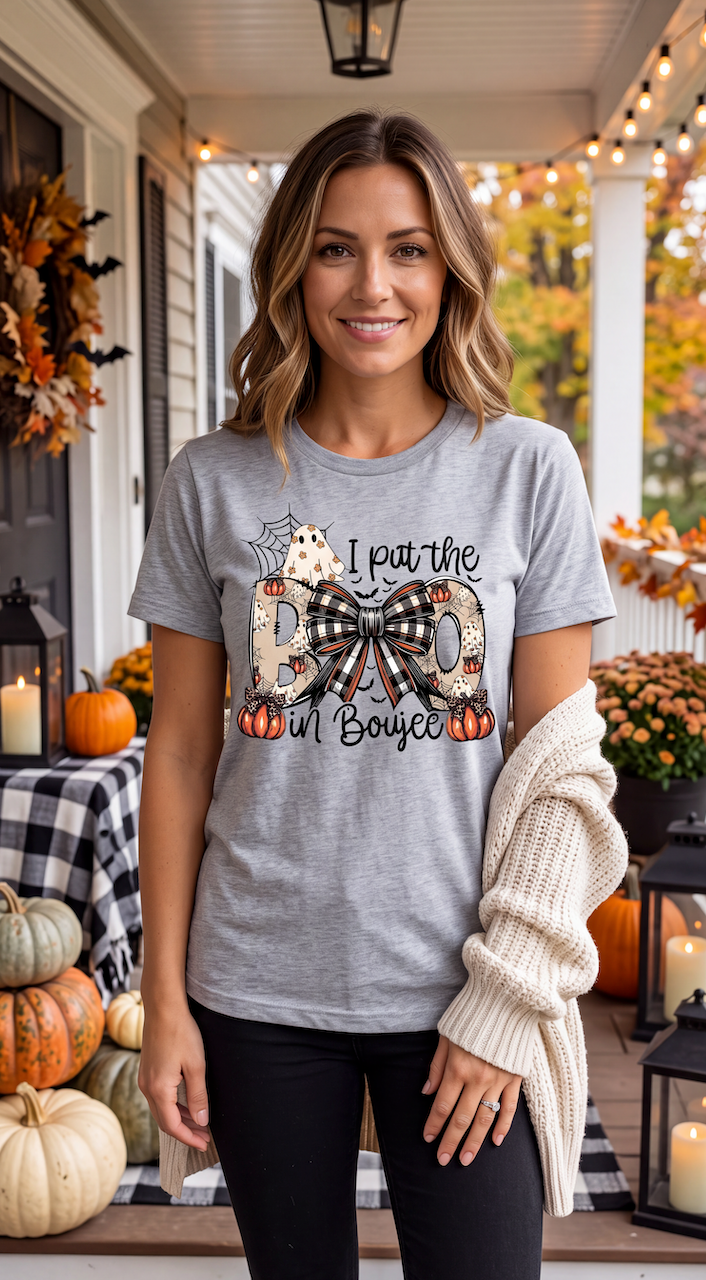 A woman stands on a decorated autumn porch wearing a heather gray graphic T-shirt with a Halloween-themed design that reads 'I put the BOO in Boyzce' with a bow and pumpkins, paired with black pants and a cream knit sweater draped over her arm.