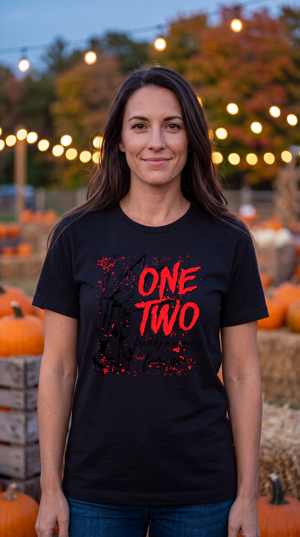 A woman wearing a black graphic t-shirt with 'ONE TWO' printed in red and black splatter-style lettering, standing in front of a blurred pumpkin patch background with warm string lights.