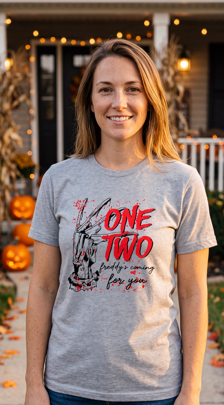 A woman wearing a heather grey graphic t-shirt with 'ONE TWO' printed in red and black splatter-style lettering, standing in front of a blurred pumpkin patch background with warm string lights.
