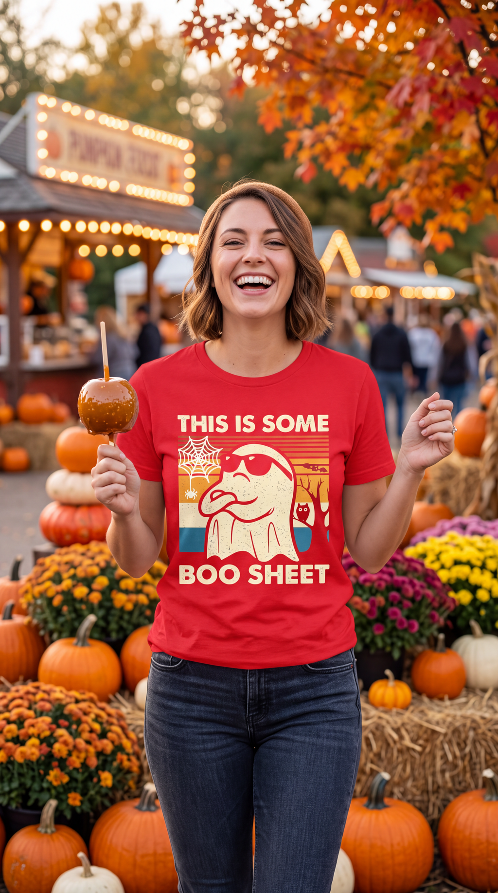 A woman wearing a red short-sleeve crewneck t-shirt featuring a retro-style graphic of a ghost wearing sunglasses with the text 'THIS IS SOME BOO SHEET' on the front, displayed on a pumpkin patch background.