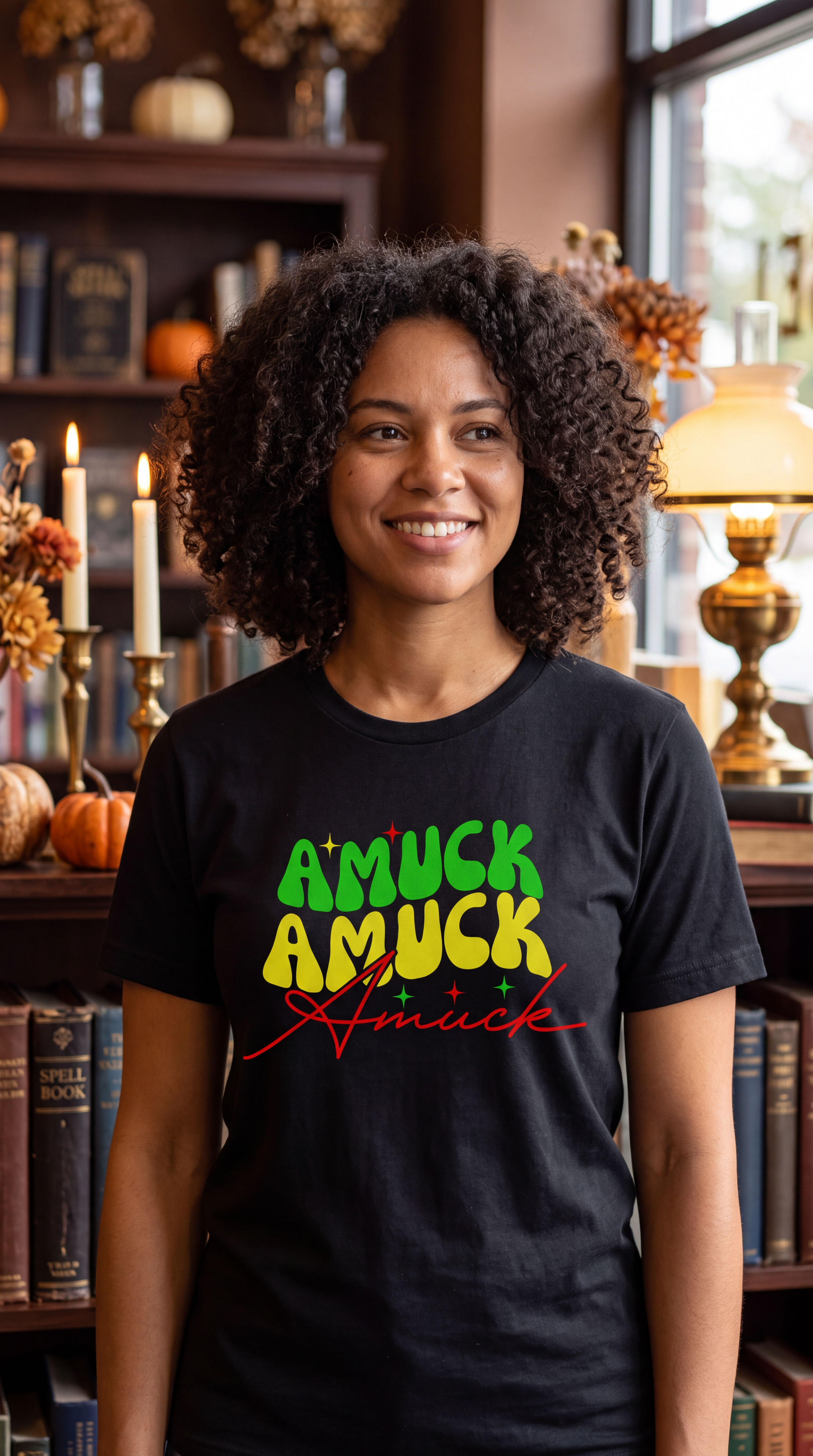 A woman wearing black short-sleeve crewneck t-shirt with a colorful graphic text print on the chest reading 'AMUCK AMUCK Amuck' in stylized fonts, shown in a library.