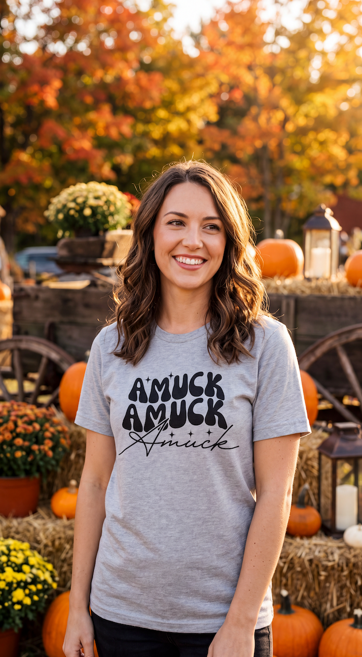 A woman wearing heather grey short-sleeve crewneck t-shirt with a black graphic text print on the chest reading 'AMUCK AMUCK Amuck' in stylized fonts, shown on a pumpkin patch.