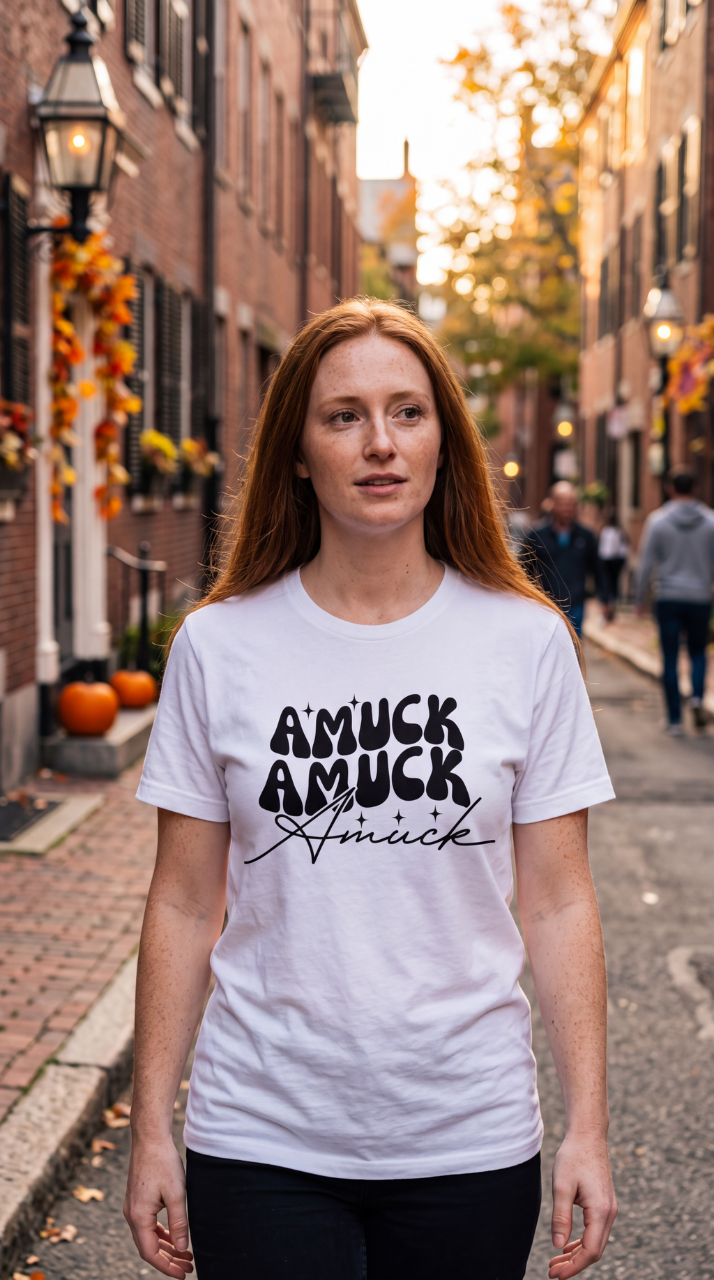 A woman wearing white short-sleeve crewneck t-shirt with a black graphic text print on the chest reading 'AMUCK AMUCK Amuck' in stylized fonts, shown on a clean street with pumpkins.