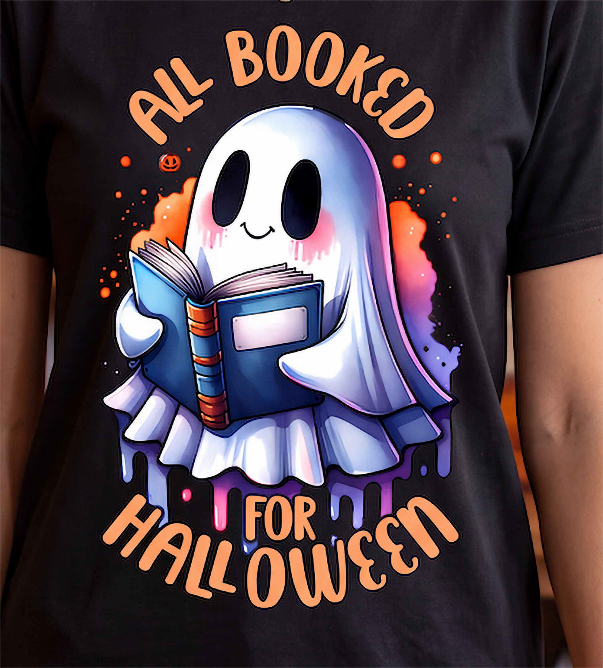 A close-up of a person wearing a black graphic T-shirt. The shirt features a cartoon ghost reading a blue book, surrounded by orange text that reads 'ALL BOOKED FOR HALLOWEEN' and colorful paint-splatter accents.