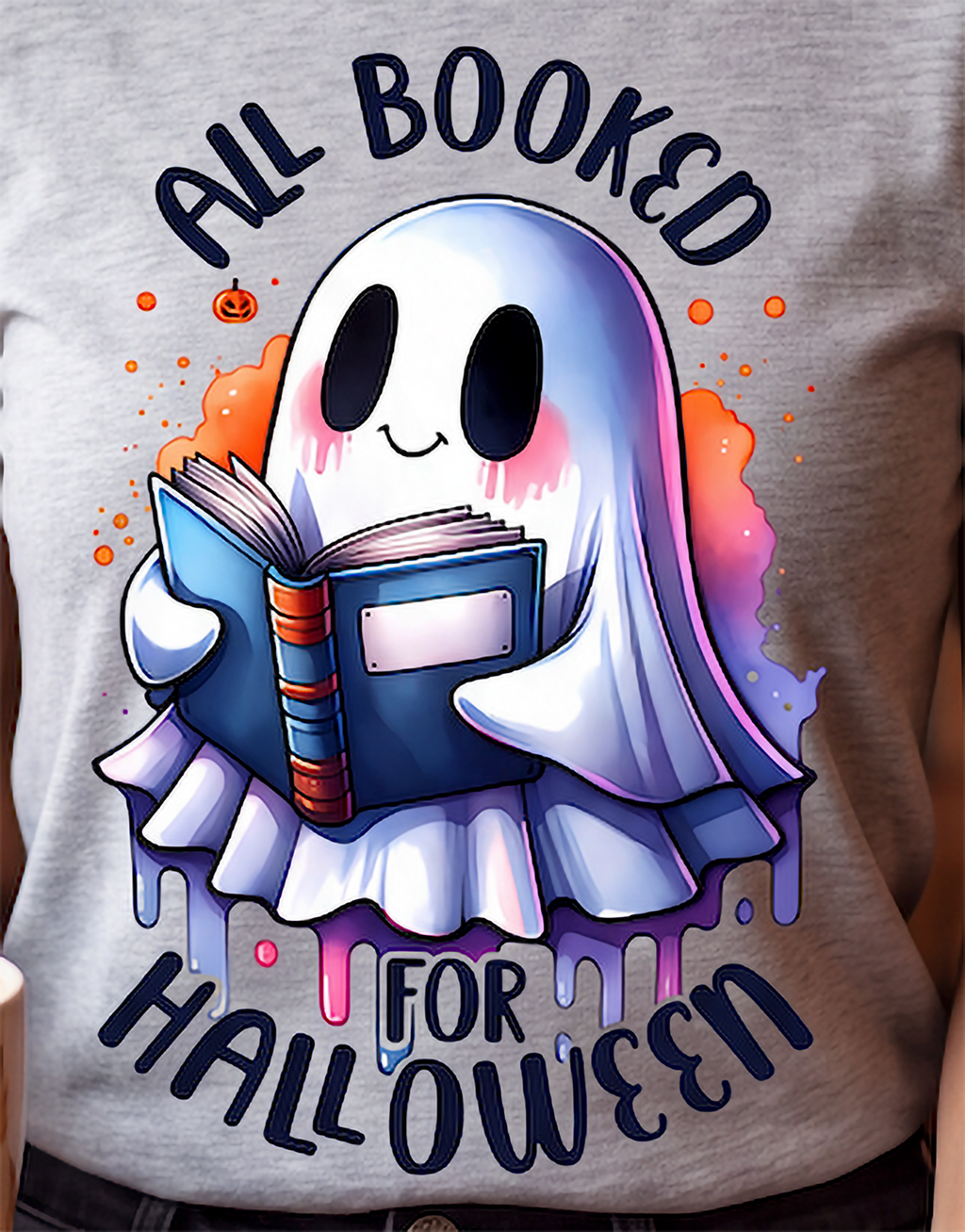 A close-up of a person wearing a heather gray t-shirt with a colorful graphic print of a cute ghost reading a book, with the text 'ALL BOOKED FOR HALLOWEEN'.