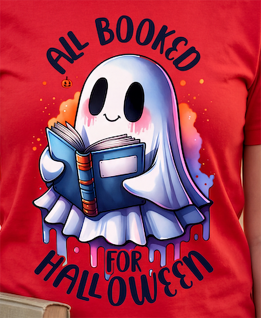 A close-up of a red T-shirt featuring a cartoon ghost graphic reading 'ALL BOOKED FOR HALLOWEEN' while holding an open book. The shirt is shown on a person, with the torso and part of the arm visible.