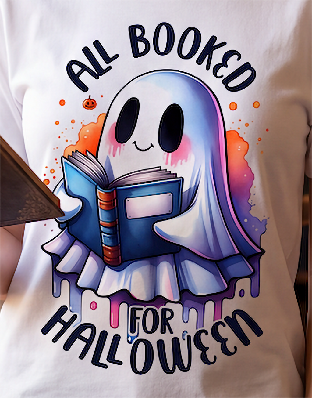 A close-up of a white T-shirt featuring a colorful graphic of a ghost reading a blue book, with the text 'ALL BOOKED FOR HALLOWEEN' in dark lettering.