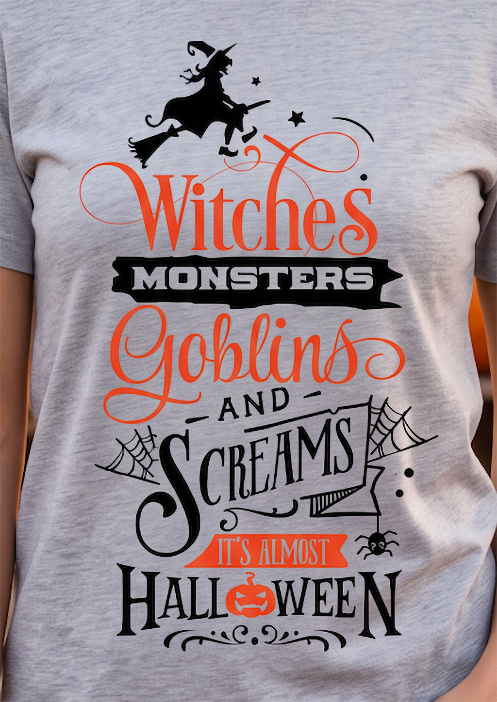 A close-up of a heather gray t-shirt featuring a Halloween graphic print with the text 'Witches Monsters Goblins and Screams It's Almost Halloween' in orange and black, along with illustrations of a witch on a broomstick, goblins, spiders, and a pumpkin.