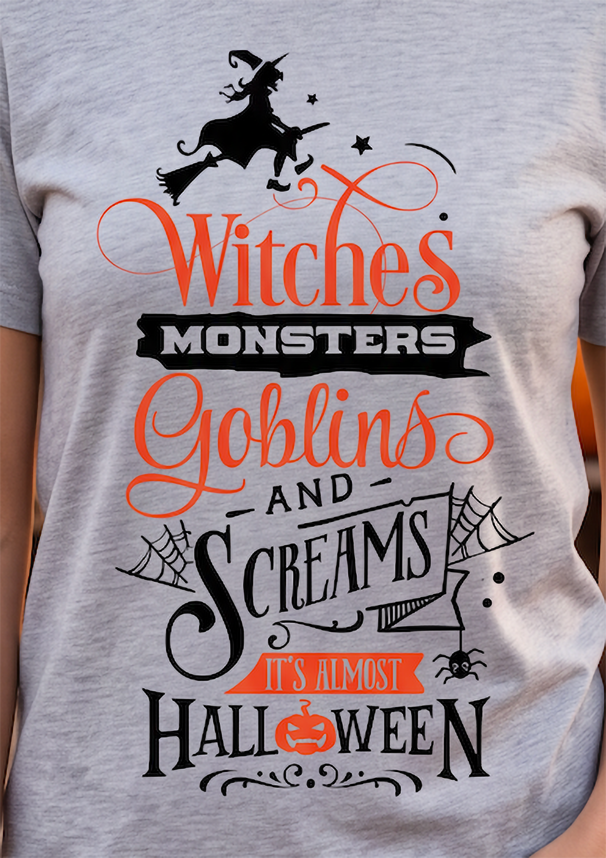A close-up of a heather gray t-shirt featuring a Halloween graphic print with the text 'Witches Monsters Goblins and Screams It's Almost Halloween' in orange and black, along with illustrations of a witch on a broomstick, goblins, spiders, and a pumpkin.