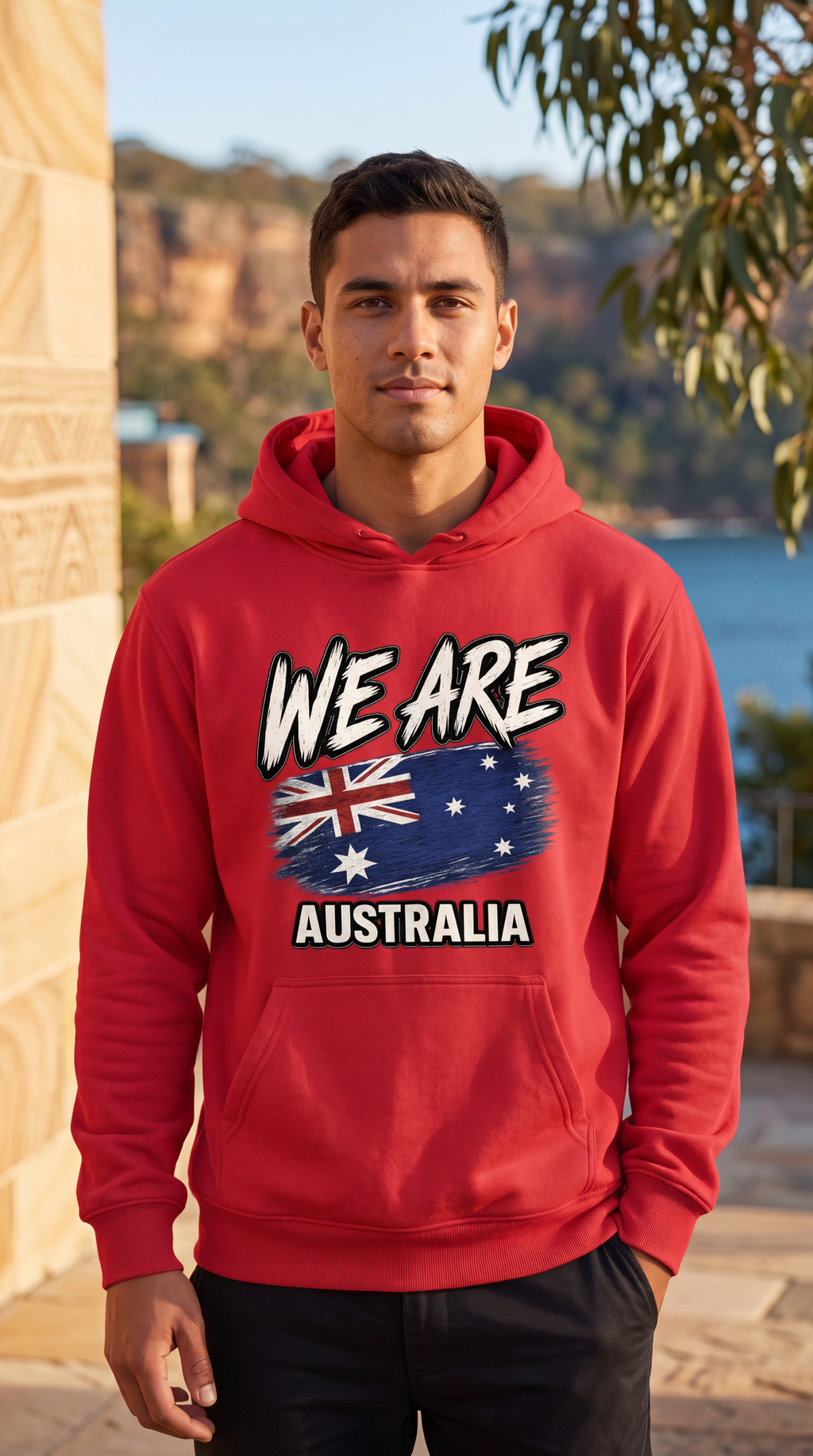 A man wearing a red hoodie with a graphic that reads 'WE ARE AUSTRALIA' and features the Australian flag, standing outdoors in front of a scenic coastal background.