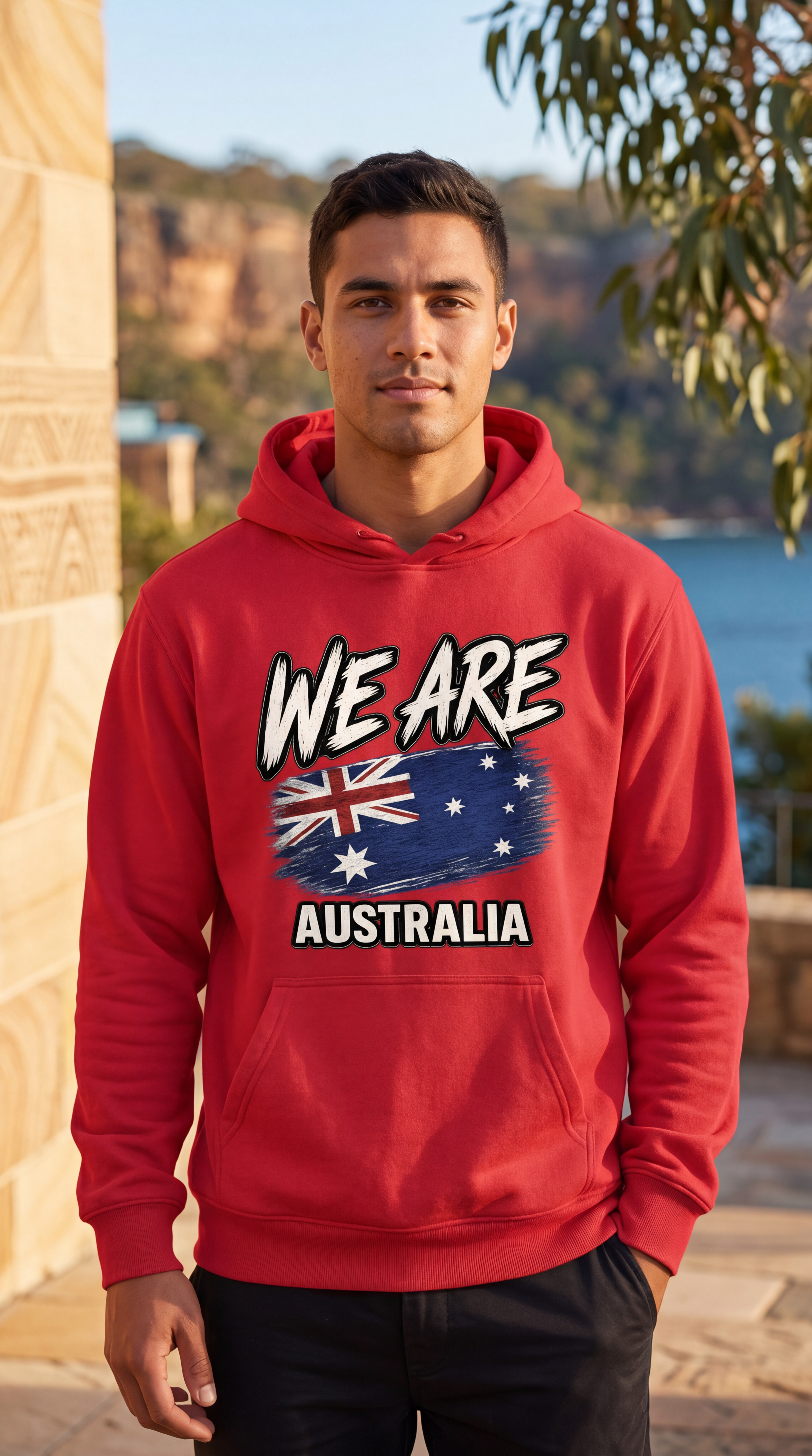 A man wearing a red hoodie with a graphic that reads 'WE ARE AUSTRALIA' and features the Australian flag, standing outdoors in front of a scenic coastal background.