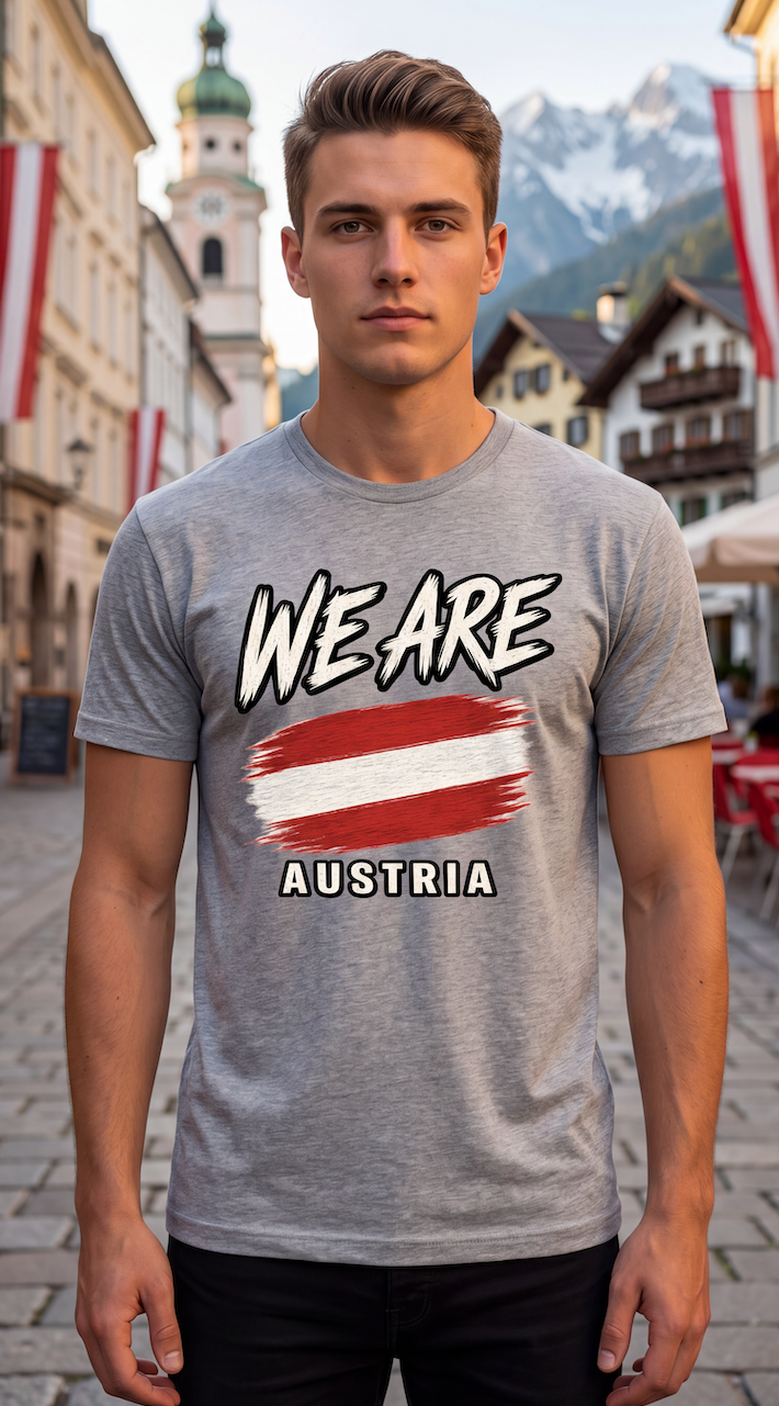 A man wearing a heather gray t-shirt with a graphic that reads 'WE ARE' above an Austrian flag and 'AUSTRIA' below, standing in an outdoor street setting with mountains in the background.