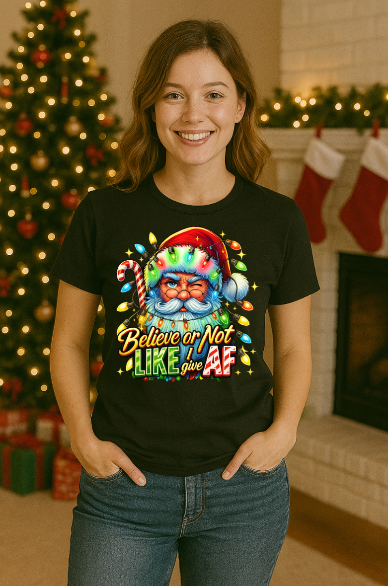 Woman wearing a black Christmas t-shirt featuring a colorful Santa Claus graphic with holiday lights and the phrase “Believe or Not I Like Give AF,” standing in a festive room with a decorated Christmas tree and stockings.