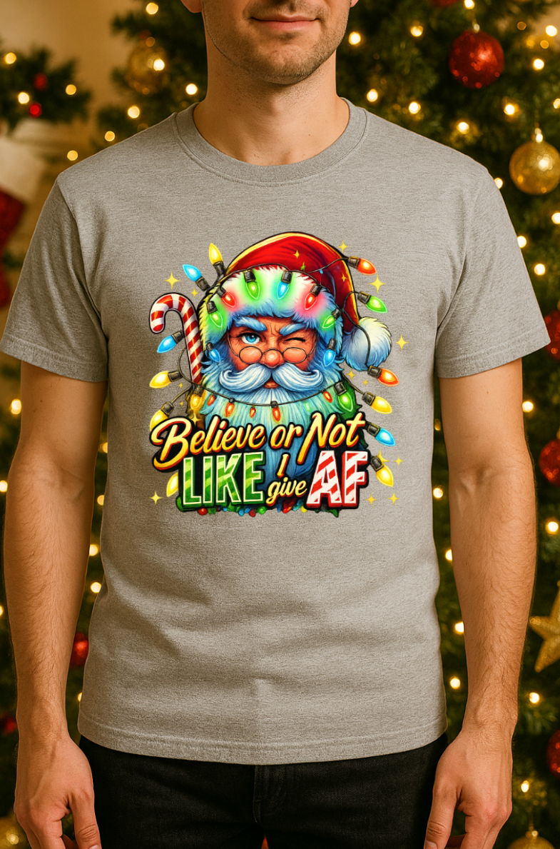 Man wearing a gray Christmas t-shirt featuring a colorful Santa Claus graphic wrapped in holiday lights with the phrase “Believe or Not I Like Give AF,” standing in front of a decorated Christmas tree.