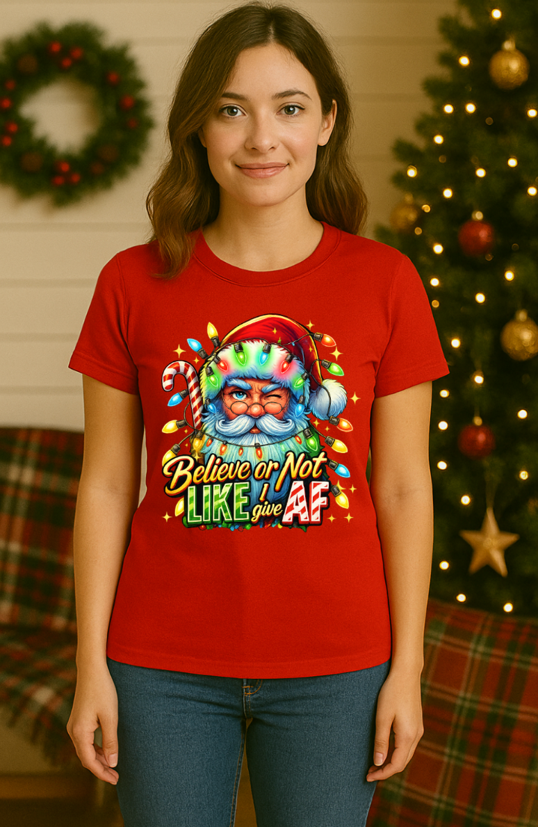 Woman wearing a red Christmas t-shirt featuring a colorful Santa Claus graphic with holiday lights and the phrase “Believe or Not I Like Give AF,” standing in a festive room with a Christmas tree and decorations.