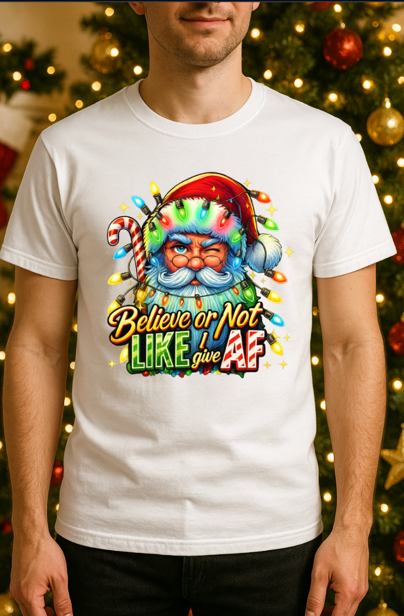 Man wearing a white Christmas t-shirt featuring a colorful Santa Claus graphic wrapped in holiday lights with the phrase “Believe or Not I Like Give AF,” standing in front of a decorated Christmas tree.