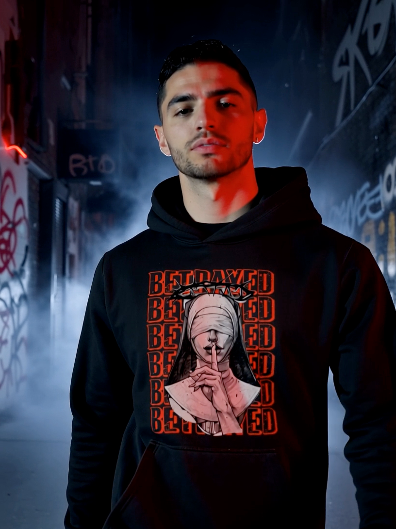 Man wearing black hoodie with “Betrayed” graphic and dark streetwear design in urban alley