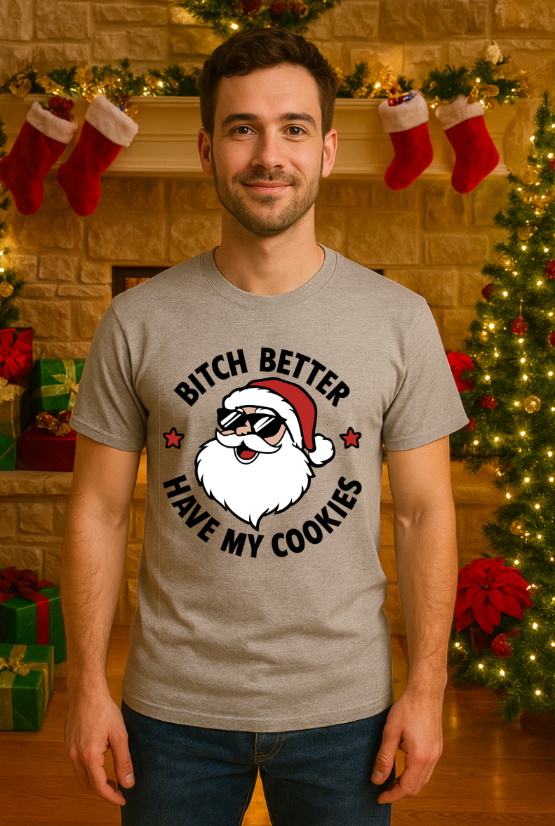 Man wearing a gray Christmas t-shirt featuring a cartoon Santa with sunglasses and a humorous cookie-themed holiday design, standing in a festive indoor setting with stockings and a decorated tree.